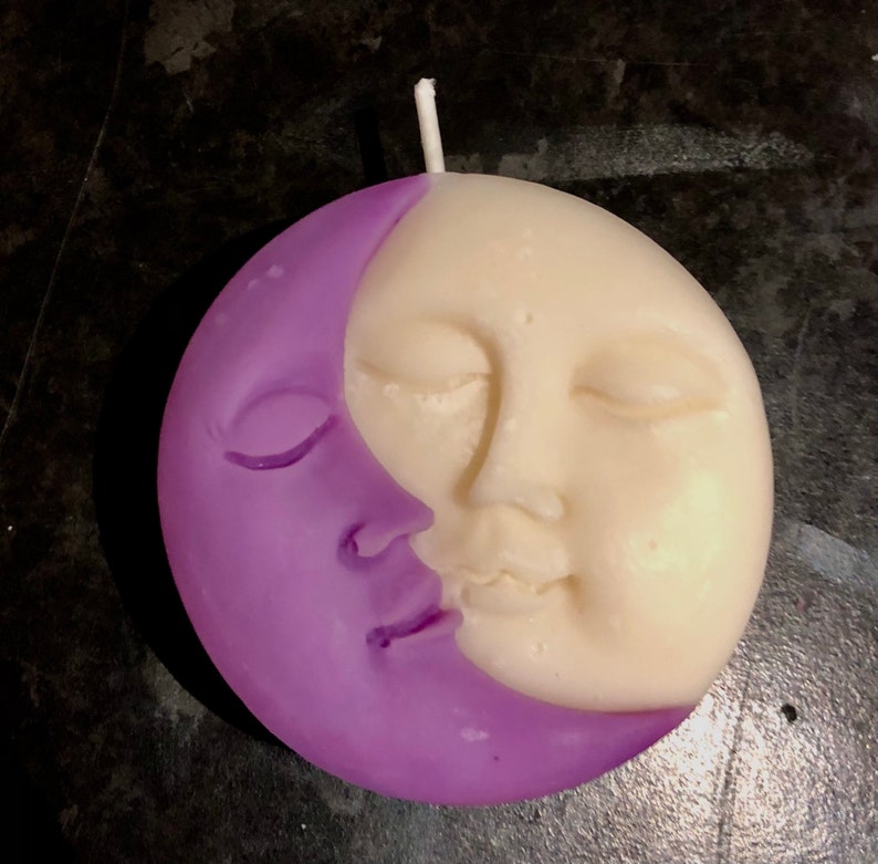 K&ouml;nnte beinhalten: Eine violette und wei&szlig;e Kerze in Form einer Mondsichel und eines Gesichts. Der Mond befindet sich auf der linken Seite der Kerze und das Gesicht auf der rechten Seite. Das Gesicht hat geschlossene Augen und einen l&auml;chelnden Mund.