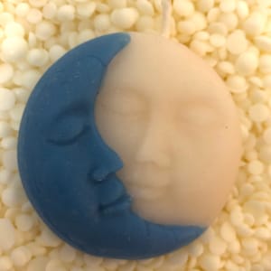 K&ouml;nnte beinhalten: Eine blaue und wei&szlig;e Kerze in Form eines Halbmondes mit einem Gesicht. Die Kerze liegt auf einem Bett aus wei&szlig;en Wachsflocken.