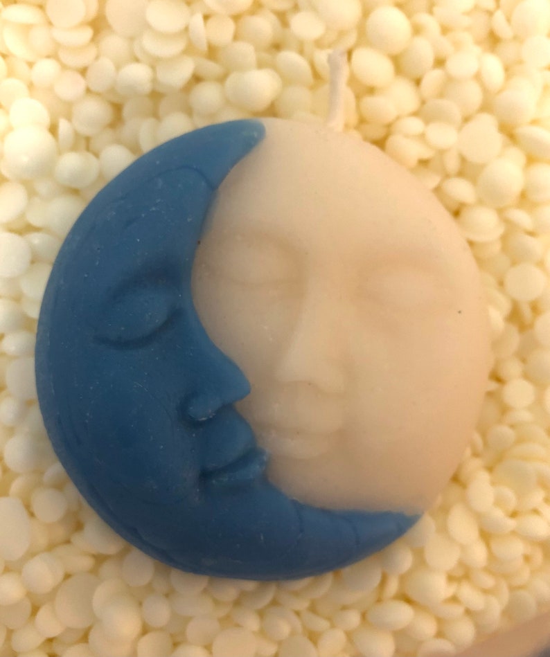 K&ouml;nnte beinhalten: Eine blaue und wei&szlig;e Kerze in Form eines Halbmondes mit einem Gesicht. Die Kerze liegt auf einem Bett aus wei&szlig;en Wachsflocken.