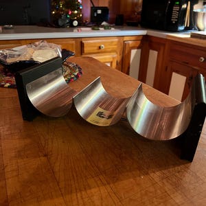May include: A set of three stainless steel and black metal kitchen tools, possibly for food preparation, arranged on a wooden surface. The tools have a curved, half-moon shape with a shiny, reflective surface. The background includes a Christmas tree and kitchen appliances.