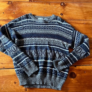 May include: A blue, gray, and white striped sweater with a geometric pattern. The sweater has a crew neck and long sleeves.