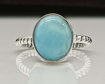 May include: A silver ring featuring a large, oval-shaped, light blue gemstone. The band has a twisted rope design. The ring is set against a white and black background, highlighting the gemstone's color and the ring's details.