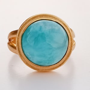 Könnte beinhalten: Ein goldfarbener Ring mit einem großen, runden, hellblauen Edelstein. Der Edelstein ist in einer goldenen Lünette gefasst und hat ein marmoriertes Aussehen. Der Ring hat ein geteiltes Schaftdesign.