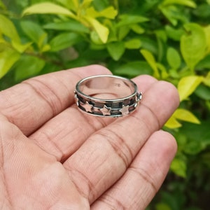 Puede incluir: Un anillo de plata con una banda de estrellas alrededor del centro. El anillo está en una mano, con un fondo verde.