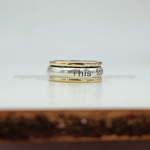 K&ouml;nnte beinhalten: Ein drehbarer Ring aus Silber und Gold mit der Gravur "This too" auf dem Band.