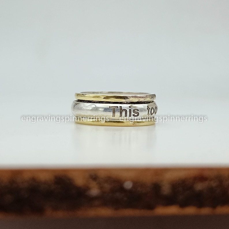 K&ouml;nnte beinhalten: Ein drehbarer Ring aus Silber und Gold mit der Gravur "This too" auf dem Band.