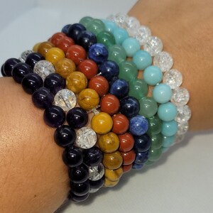 Op de afbeelding: Een stapel van zeven kleurrijke kralenarmbanden, elk met een andere edelsteen. De armbanden zijn gemaakt van amethyst, tijgeroog, rode jaspis, sodaliet, aventurijn, turquoise en helder kwarts.
