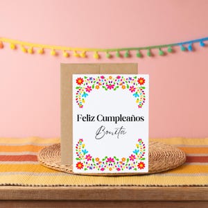 May include: A white card with a colourful floral border and the text "Feliz Cumpleaños Bonita" in black and white.