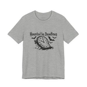 Peut inclure: Un t-shirt gris chiné à manches courtes avec une impression graphique noire représentant une horloge fondante avec des chiffres romains et des chauves-souris. Le texte "Haunted by Deadlines" est imprimé dans une police dentelée.
