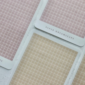 Może przedstawiać: Dwa arkusze papieru w kratkę z nadrukowanym tekstem "PAPER DREAMSCAPE" na dolnej krawędzi. Papier po lewej stronie jest jasnoróżowy, a papier po prawej stronie jest jasnobeżowy.