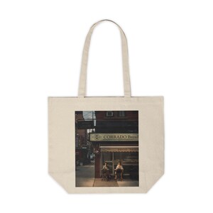 May include: A vintage photograph of a storefront with the sign "Corrado Bread" above the entrance. Two people are sitting outside the bakery, enjoying the warm weather.