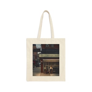 May include: A vintage photograph of a storefront in New York City. The shop is called "Corrado Bread" and has a sign that says "Bread & Sandwiches". Two people are sitting outside the shop.