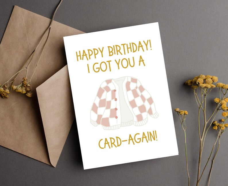 May include: A white card with gold text that reads "Happy Birthday! I got you a card-again!" and a cartoon image of a pink and white checkered cardigan.