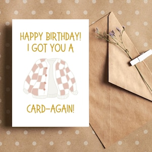 May include: A white card with the text "Happy Birthday! I got you a card-again!" in gold lettering. A pink and white checkered cardigan is illustrated in the center of the card.
