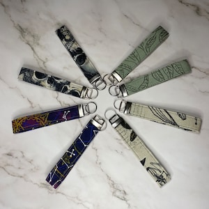 May include: An assortment of key fobs featuring diverse fabric patterns, such as skulls, leaves, and celestial motifs. Each fob is fitted with a silver-toned metal ring. The key fobs are arranged in a circular design on a marble surface.