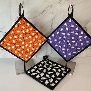May include: Three quilted pot holders with a ghost and crescent moon pattern. The pot holders are orange, purple, and black.