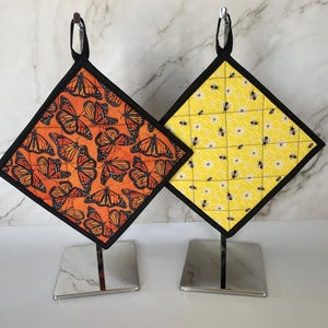 May include: Two quilted pot holders with black trim. One pot holder features a yellow background with black bees and white flowers. The other pot holder features an orange background with black butterflies.