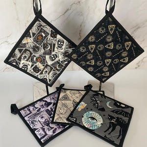 May include: Set of four black and white kitchen pot holders with a spooky Halloween theme. The pot holders feature various designs including an eye, a skull, a cat, a bat, and a crescent moon. The pot holders are made of fabric and have a black border.
