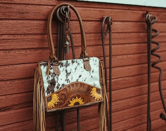 Könnte beinhalten: Eine braune Ledertasche mit Fransenbesatz und einem Sonnenblumenmuster. Die Tasche hängt an einem Metallhaken an einer roten Holzwand.