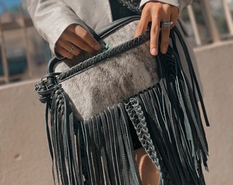 May include: A black and white cowhide handbag with a long black fringe and a braided strap. The bag is being held by a person wearing a white blazer.