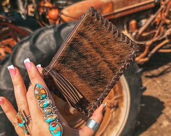 Op de afbeelding: Een bruine leren portemonnee met een kwastje. De portemonnee wordt vastgehouden in een hand met turquoise en zilveren ringen. De hand draagt ​​verschillende zilveren armbanden met turquoise stenen en gegraveerde tekst.