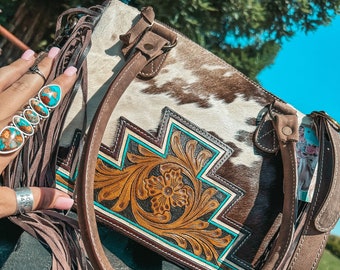 Könnte beinhalten: Eine braune und weiße Kuhfell-Handtasche mit türkisfarbenen und braunen Lederelementen. Die Tasche hat einen Fransenbesatz und einen oberen Henkel.