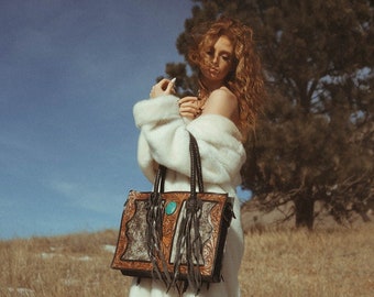 May include: A brown and black leather tote bag with intricate floral tooling, turquoise accents, and fringe detailing. The bag has braided handles and is held in a field of tall, dry grass under a blue sky.