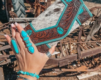 May include: A brown leather clutch with turquoise accents and a cowhide pattern. The clutch is held in a hand with turquoise rings and bracelets.