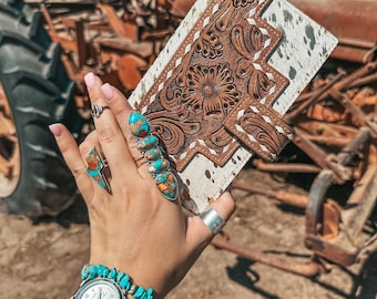 May include: A brown leather wallet with a white and brown cowhide pattern. The wallet has intricate floral designs and stitching. The wallet is being held by a hand wearing turquoise jewelry and a silver watch.