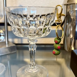 May include: A clear crystal coupe glass with a gold-colored keychain featuring three green olive beads with red centers. The glass has a diamond pattern and a long stem. A cocktail shaker is visible in the background.