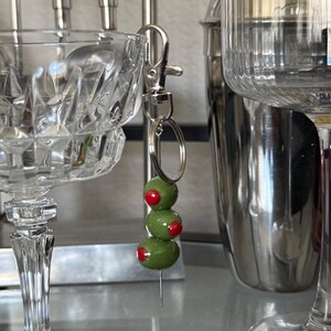 May include: A keychain featuring three green olive beads with red pimento centers. The olives are strung on a silver-colored pin and attached to a silver clasp and keyring. The background includes a cocktail glass and a stainless steel cocktail shaker.