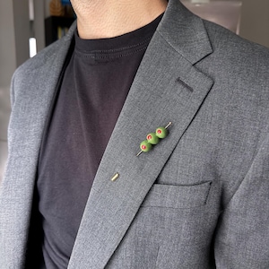May include: A lapel pin featuring three green olives with red pimentos skewered on a thin gold-toned metal needle. The pin is attached to the left lapel of a charcoal gray blazer worn over a plain black t-shirt, adding a distinct accessory detail.