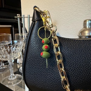 May include: A black leather handbag with a gold chain strap and a gold-tone clasp. A charm with three green olives with red pimentos on a toothpick hangs from the bag's hardware. The bag is branded with the name "MICHAEL".