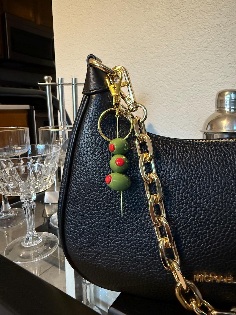 May include: A black leather handbag with a gold chain strap and a gold-tone clasp. A charm with three green olives with red pimentos on a toothpick hangs from the bag's hardware. The bag is branded with the name "MICHAEL".
