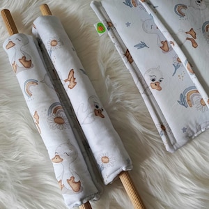 May include: Baby bedding set with a white and light grey duck and daisy pattern. The set includes two cylindrical bumpers with wooden dowels and a rectangular pad. The fabric has a soft, plush appearance. The measurements are in centimetres.