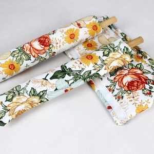 Könnte beinhalten: Blumen-Nudelholzbezüge und ein passender quadratischer Topflappen. Der Stoff hat einen weißen Hintergrund mit großen orangefarbenen, gelben und grünen Blumenmustern. Die Nudelrollen sind aus Holz.