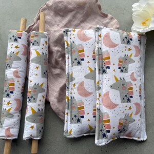 May include: White fabric items featuring a unicorn, moon, and star design. Two cylindrical items are on wooden dowels. Two rectangular items are also visible. A pink cloth and a white flower are in the background.