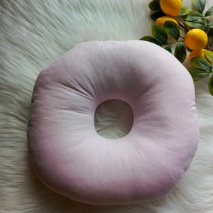 May include: A pale pink, donut-shaped pillow with a central hole. The pillow is set on a white, fluffy surface, with a sprig of lemons in the background. The pillow is designed for comfort and support.