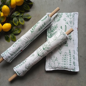 Puede incluir: Un juego de textiles de cocina blancos con estampado botánico verde. El juego incluye dos fundas para rodillo y una almohadilla rectangular. Las fundas para rodillo tienen mangos de madera. Una rama de limones amarillos está en el fondo.
