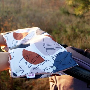 Può includere: Un paio di copri manici per passeggino con sfondo bianco e un motivo astratto di foglie colorate. Le coperture sono realizzate in tessuto morbido e hanno una chiusura a bottone. Il testo "Sleepy" è visibile nell'angolo in basso a destra della copertura.
