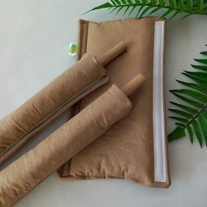 Op de afbeelding: Twee bruine deurstoppers van stof met houten pinnen erin. De deurstoppers zijn bevestigd aan een bruin stoffen kussen met een witte rits en witte klittenband.