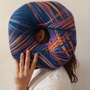 May include: A circular cushion with a central aperture, adorned with a pattern of orange and purple leaf motifs against a blue backdrop. The cushion is held in front of a person's face.