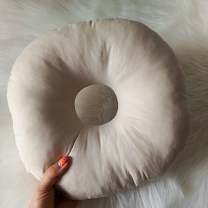 May include: A beige donut-shaped pillow with a central hole. The pillow is made of a soft, plush material. The pillow is held by a hand with orange and black nail art. The pillow is on a white, fluffy surface.