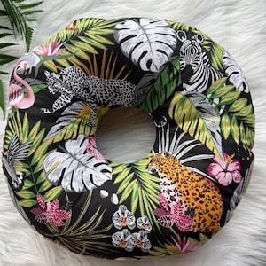 May include: A circular cushion with a black background and a colourful tropical print. The design includes leopards, zebras, flamingos, and lush green foliage, including palm leaves and flowers. This cushion is a decorative home accessory.