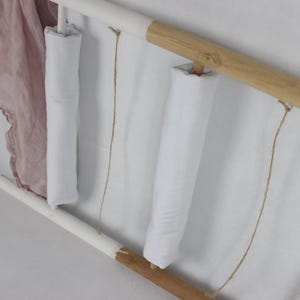 May include: Three white fabric rolls are hanging on a wooden and white frame. The rolls are tied with twine and are hanging from the frame.