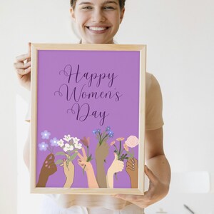 May include: A framed print with a purple background and the text "Happy Women's Day" in a purple script font.  The print features a design of multiple hands of different skin tones holding flowers.