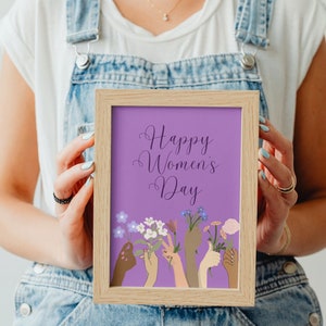 May include: A framed print with a purple background and the text "Happy Women's Day". The print features a colorful illustration of hands holding flowers.