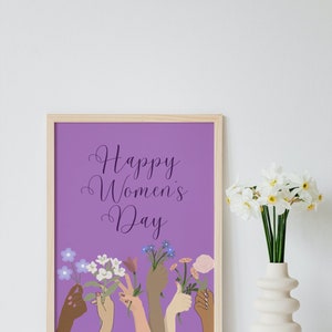 May include: A framed print with a purple background and the text "Happy Women's Day". The print features a row of diverse hands holding flowers.