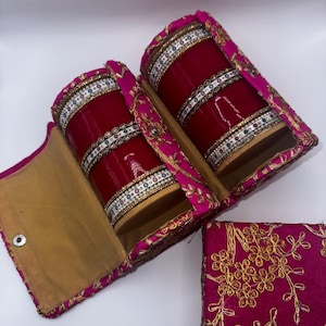 May include: Two red bangles with gold and white embellishments are displayed in a pink and gold embroidered box. The box has a hinged lid and a separate smaller box.