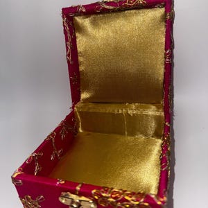 Puede incluir: Una pequeña caja de tela rosa con bordados dorados y un cierre dorado. La caja está abierta y forrada con satén dorado.